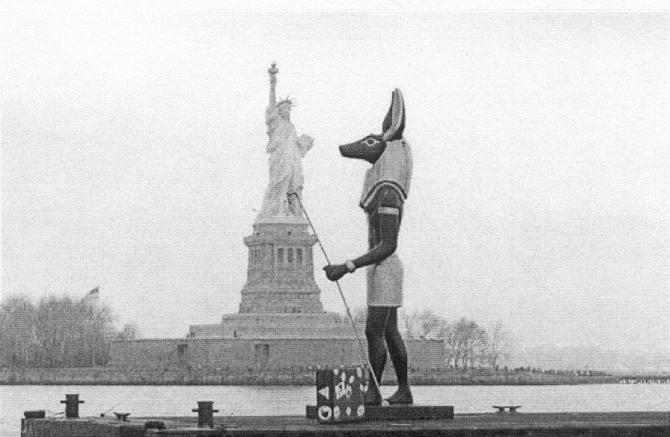 45 Anubis and the Death of America A 25-foot tall replica statue of the Egyptian god Anubis, with a suitcase at his feet, passes in front of the Statue of Liberty while taking a tour of the New York