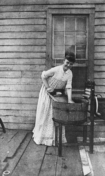 English: Woman handwashing clothes on the veranda of a house, 1902-1904 Young woman washing clothes by hand in a wooden basin on a washstand with a washboard and wringer.