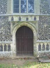 There are flushwork panels in its angled buttresses, with more above the doorway and the west window has a crocketted dripstone with finial and angel headstops.