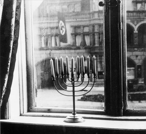 A Menorah in an apartment window overlooking Nazi Party Headquarters bedecked with its Nazi flag (1931). Kiel, Germany, in the house of Rabbi Akiva and Rachel Posner on Hanukkah, 1931.