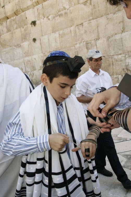 A boy putting on Tefillin after his Bar Mitzvah in Hebrew. Friends and relatives watch in the synagogue.