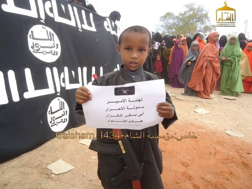 A child supporter of HSM stands besides a large ISIS/HSM banner, holding a placard that reads: Greetings to the amir in the state of free men: Abu Bakr al-